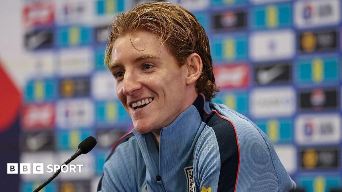 A smiling Anthony Gordon, in a blue zip-up top, sits by a microphone at an England news conference