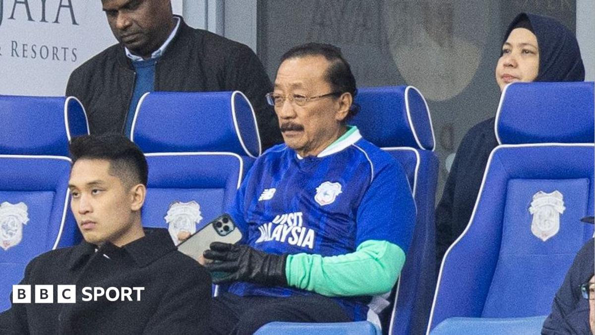 Vincent Tan wears a blue football shirt while sat in a blue seat inside stadium. He holds a mobile phone in hand.